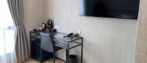 Habitación de hotel moderna con un escritorio de vidrio, una silla negra, un cubo de basura negro, un televisor de pantalla plana montado en la pared y una ventana con cortinas.
