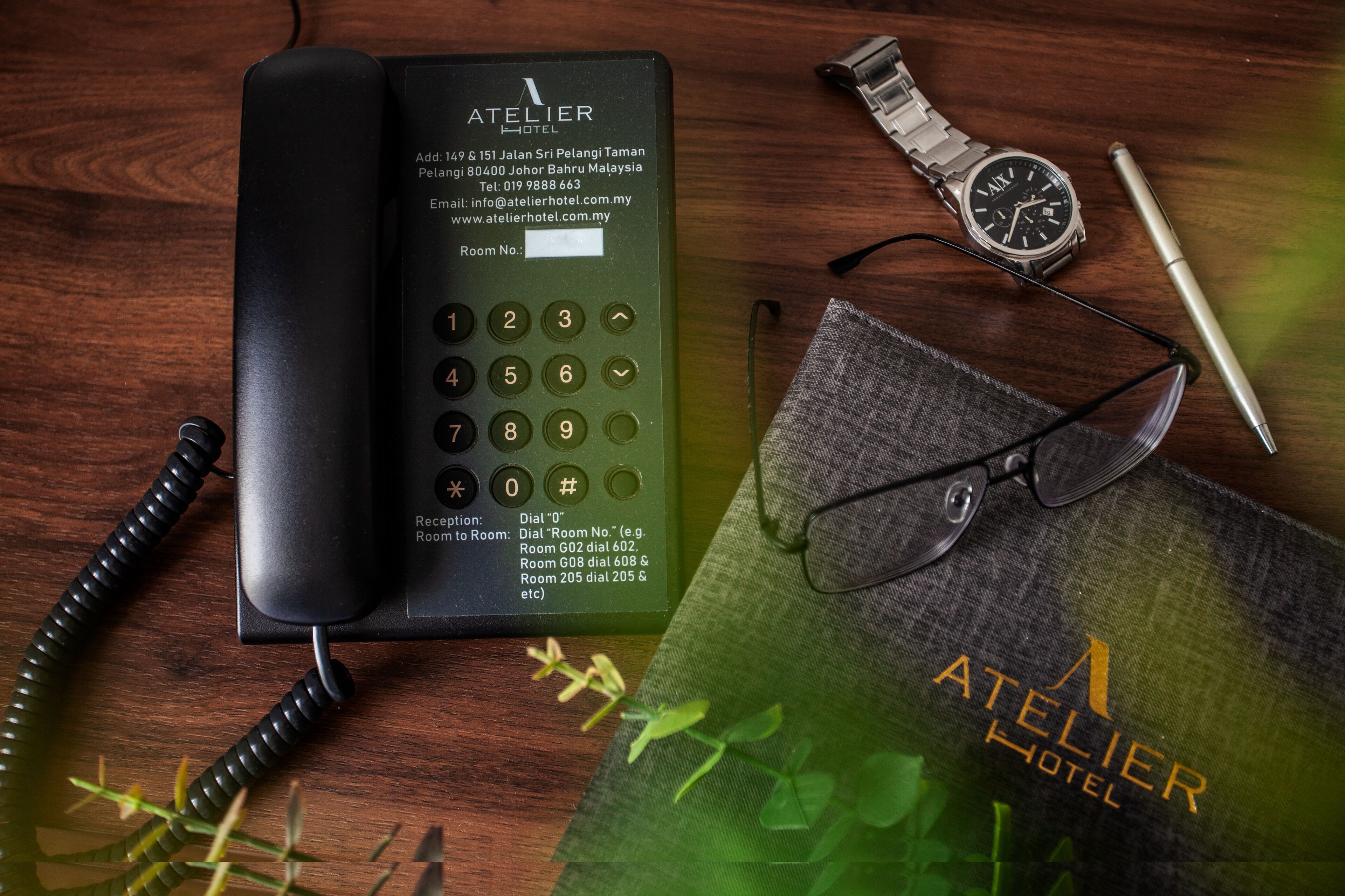 A hotel phone, a wristwatch, a pen, and a pair of glasses on a wooden surface with a plant.