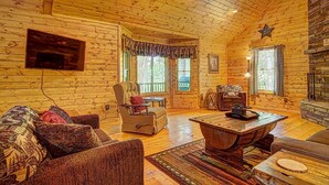 Interior acogedor de una cabaña de madera con techo de madera, chimenea, televisor montado en la pared y una zona de estar cómoda con sofá y sillones.