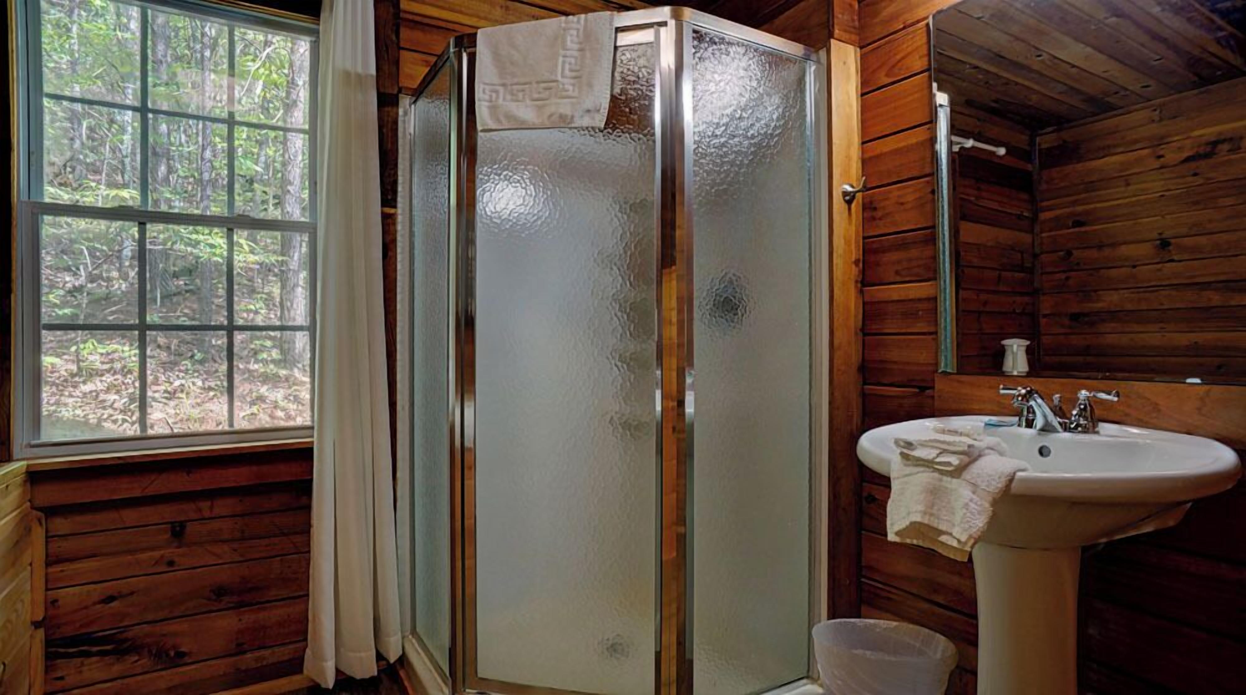 Une salle de bain rustique, avec des murs en bois, une cabine de douche en verre, un lavabo et une fenêtre agrémentée de rideaux.