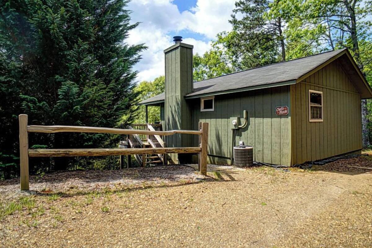 A rustic cabin with a wooden deck and a metal gate, surrounded by dense forest.