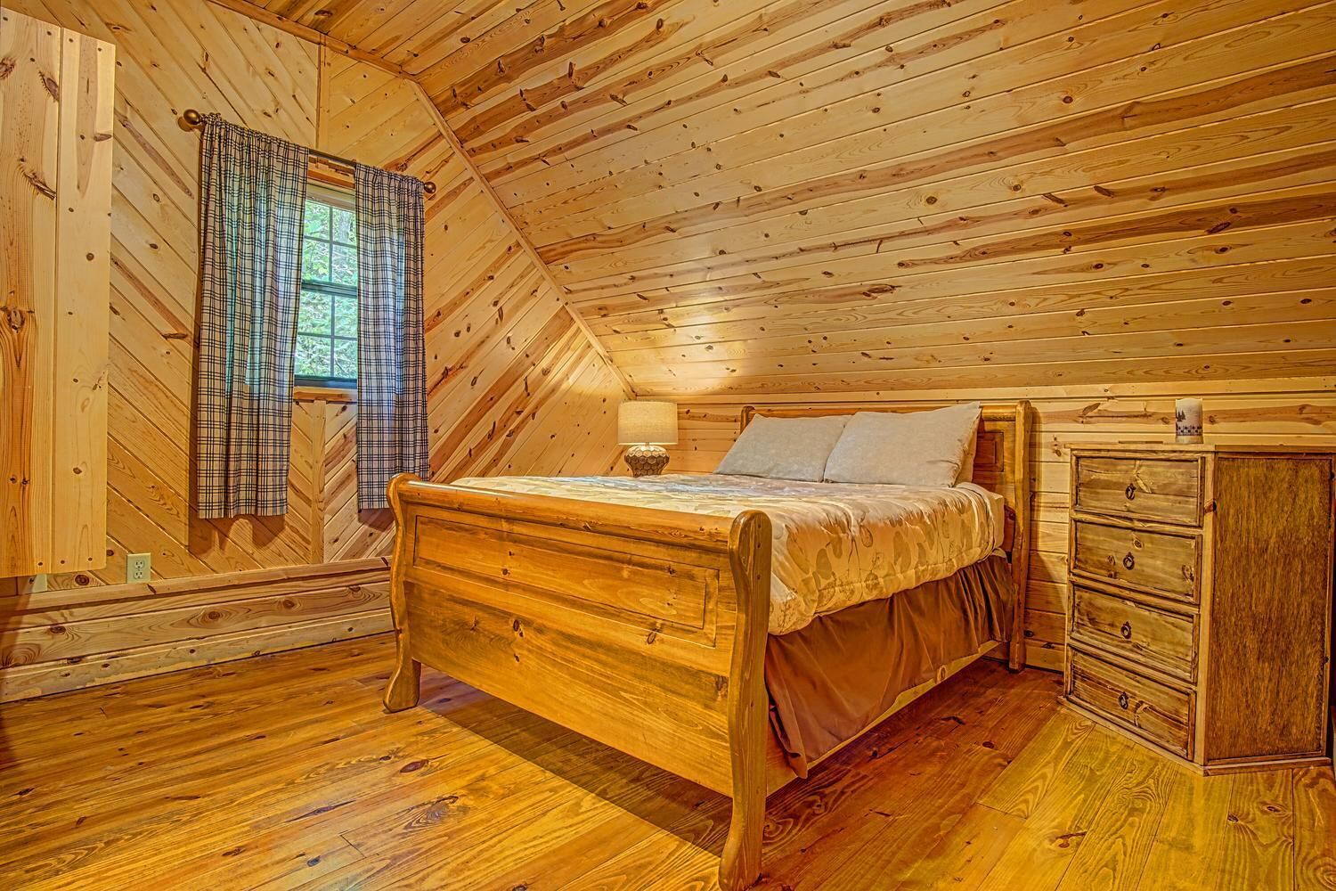 Une chambre à coucher dans un chalet en bois, avec un lit, une commode et une fenêtre ornée de rideaux à carreaux.