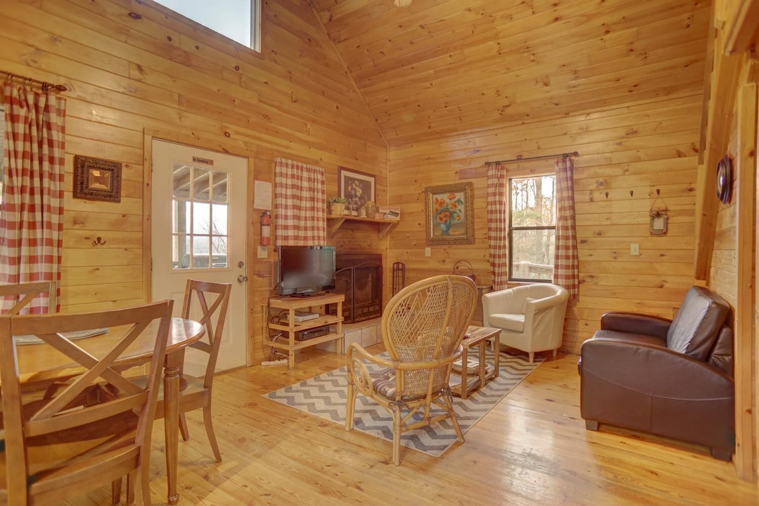 A cozy cabin interior with wooden walls, a fireplace, a TV, a dining table with chairs, a sofa, and a patterned rug.