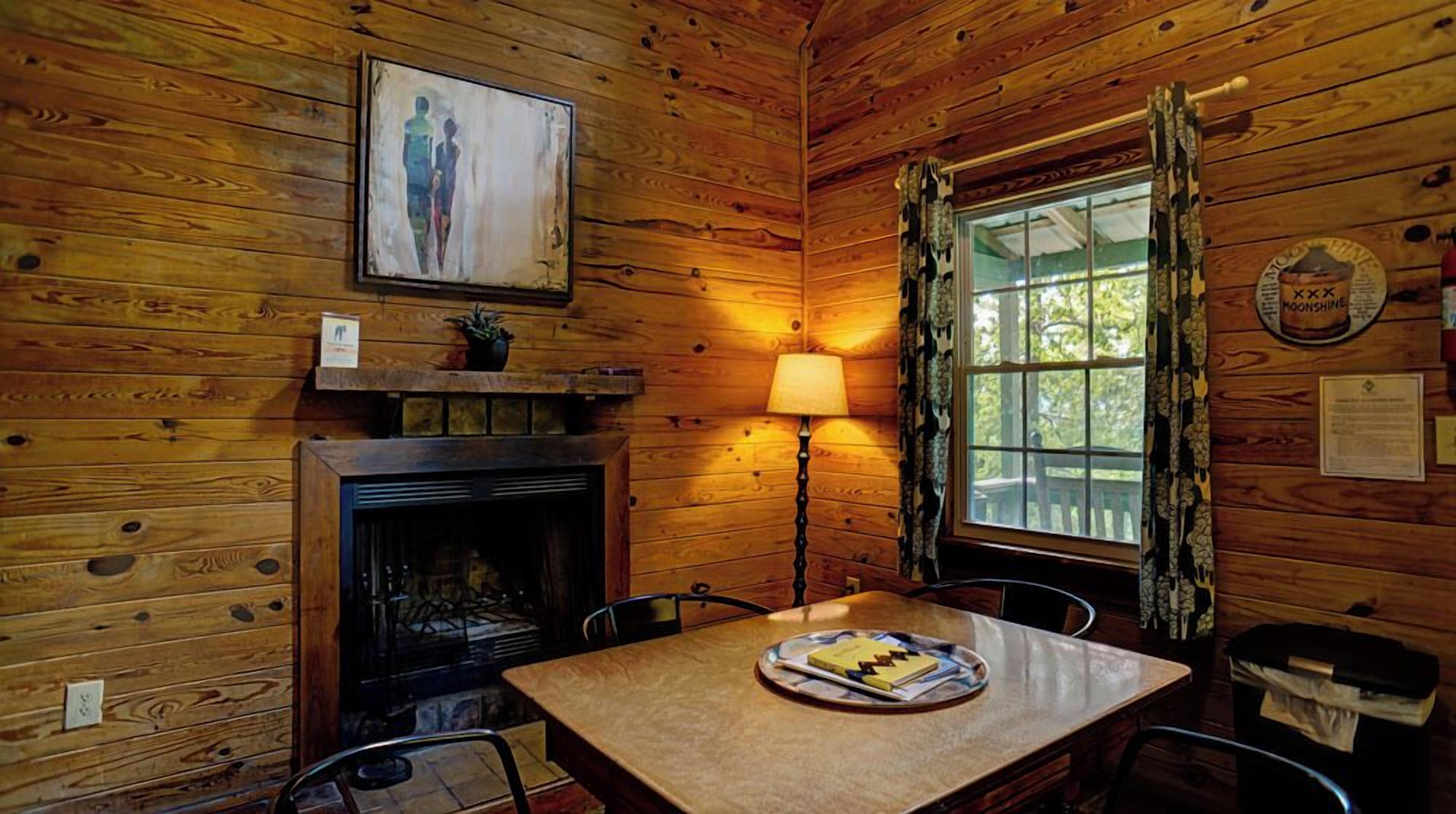 Une pièce chaleureuse dans un chalet, avec des murs en bois, une cheminée, une table sur laquelle se trouve une assiette de nourriture, une lampe et une fenêtre avec des rideaux.