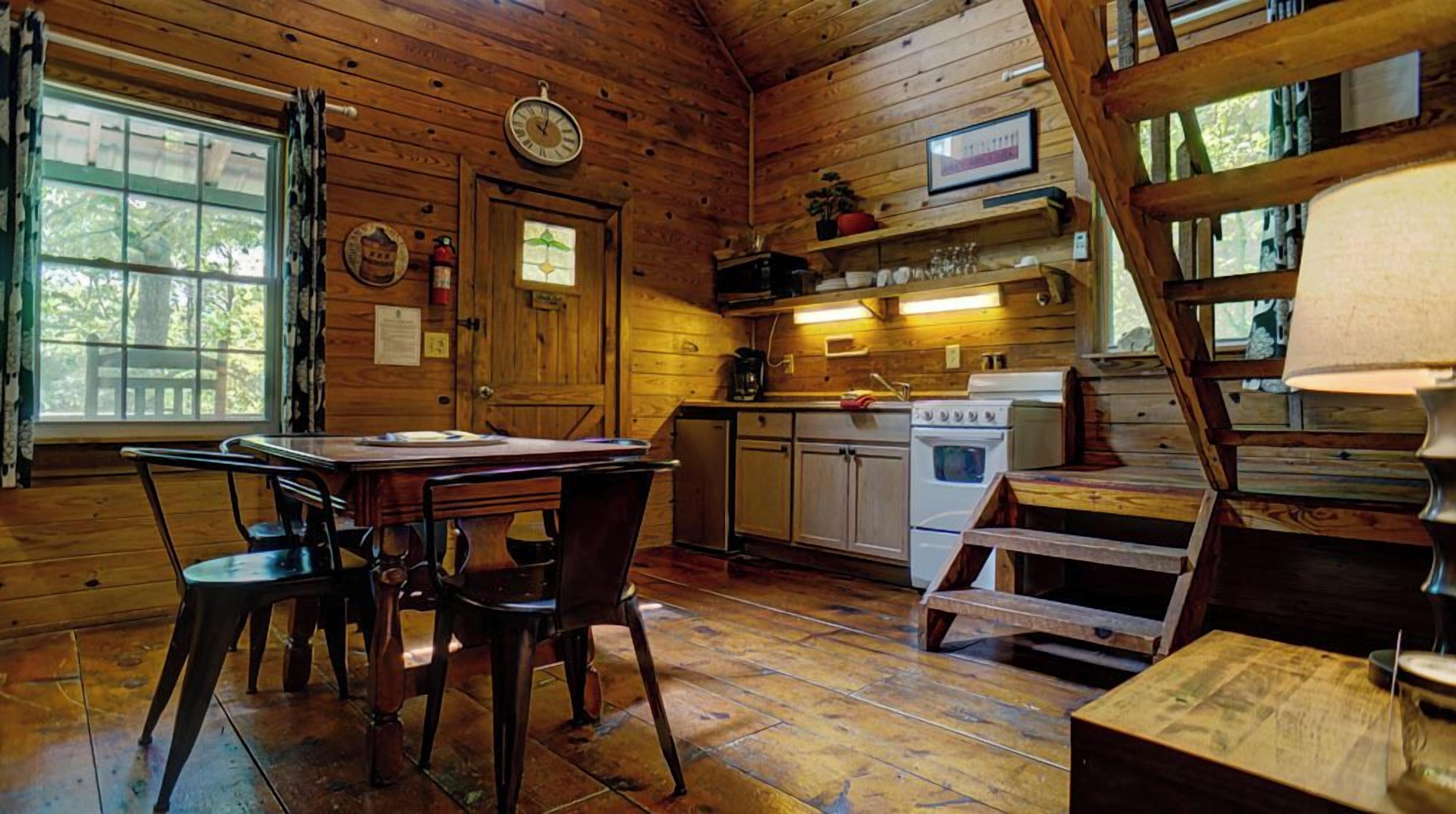 L’intérieur d’un chalet en bois rustique comprend une table à manger, des chaises, une cuisine avec un poêle et une fenêtre avec des rideaux.