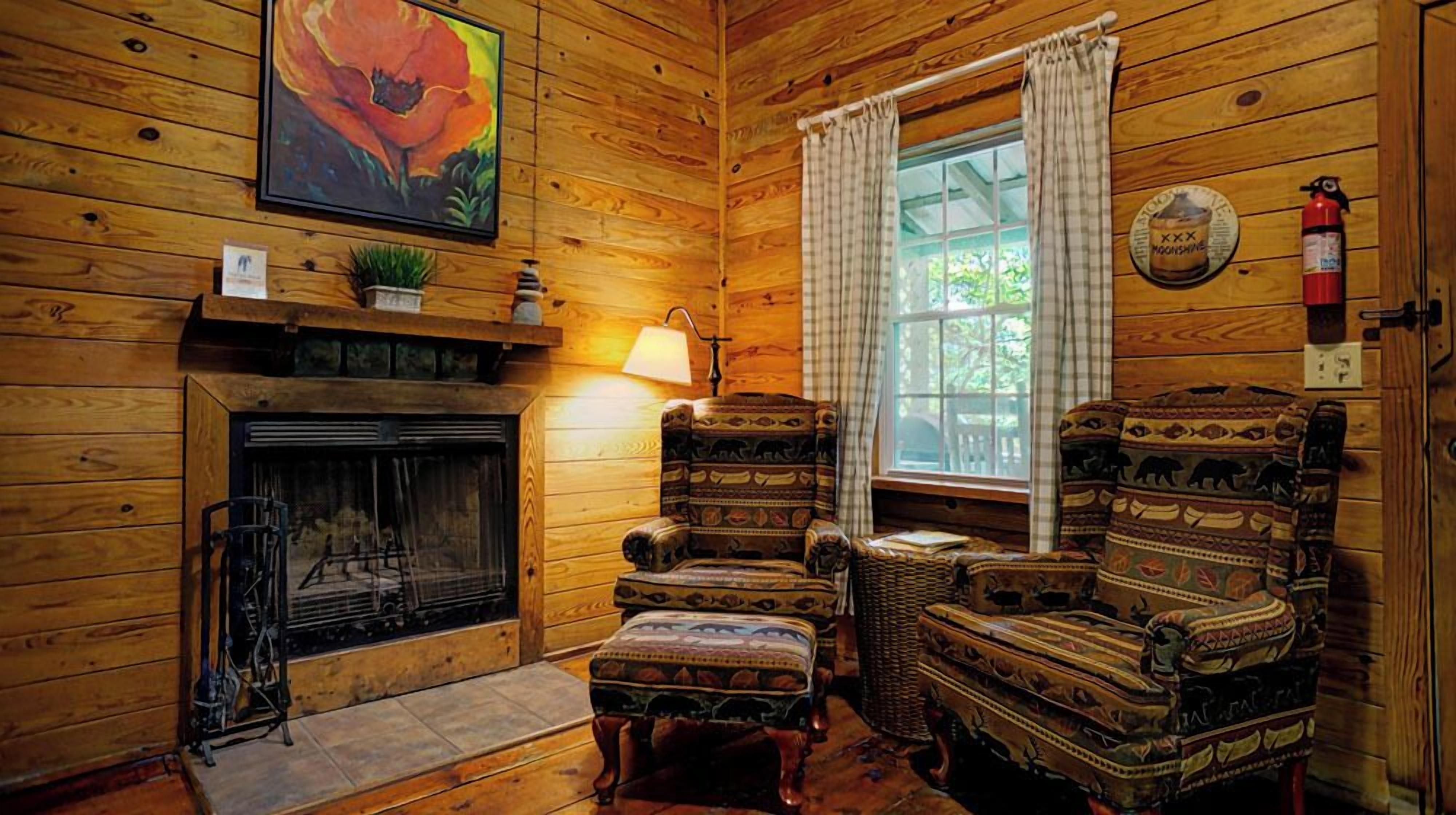 A cozy log cabin interior with a fireplace, patterned armchairs, a wicker chair, and a window with curtains.