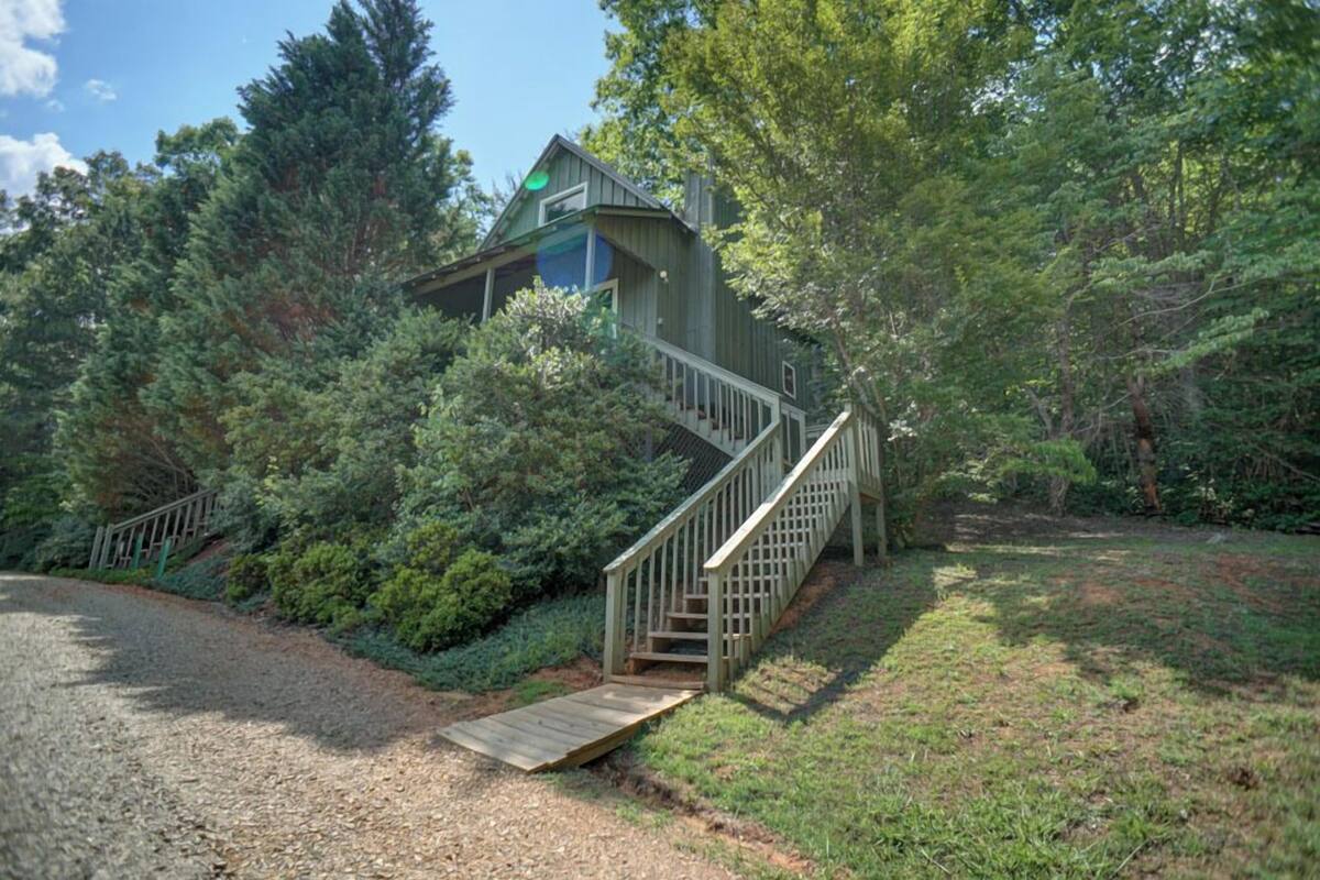 A wooden cabin with a staircase leading to a deck, surrounded by dense forest.