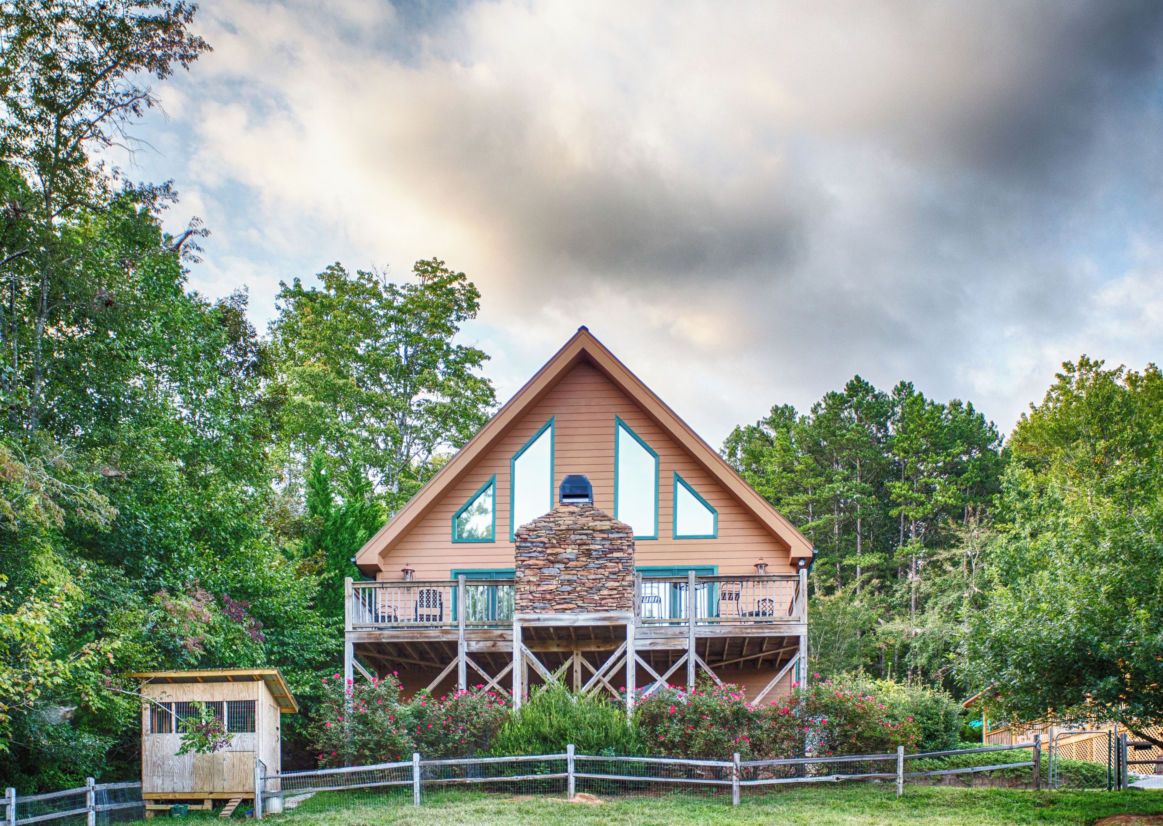 A two-story wooden house with a stone fireplace, surrounded by lush greenery and a wooden fence.