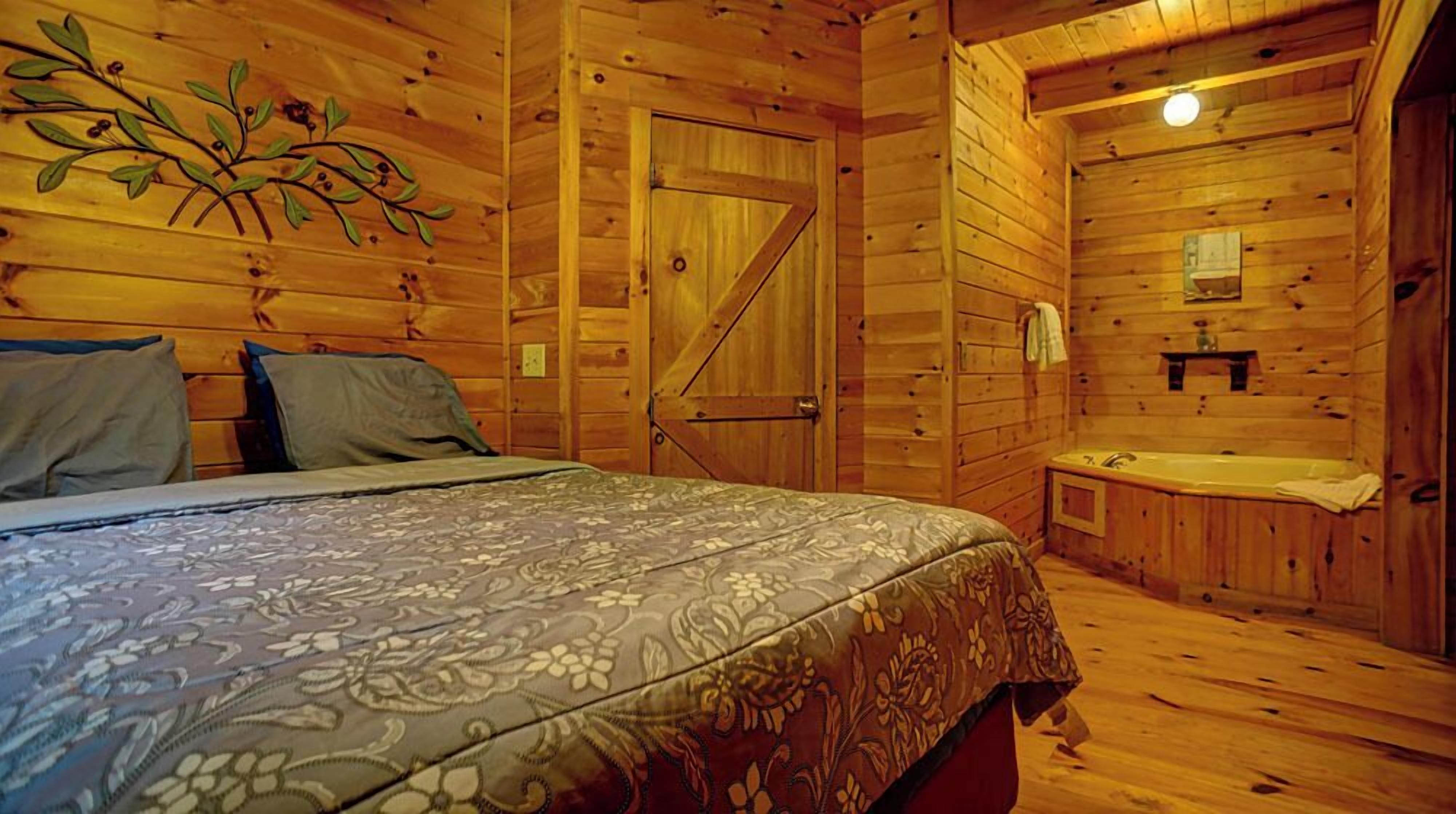 A wooden cabin room with a bed, a door, and a small bathroom with a bathtub.