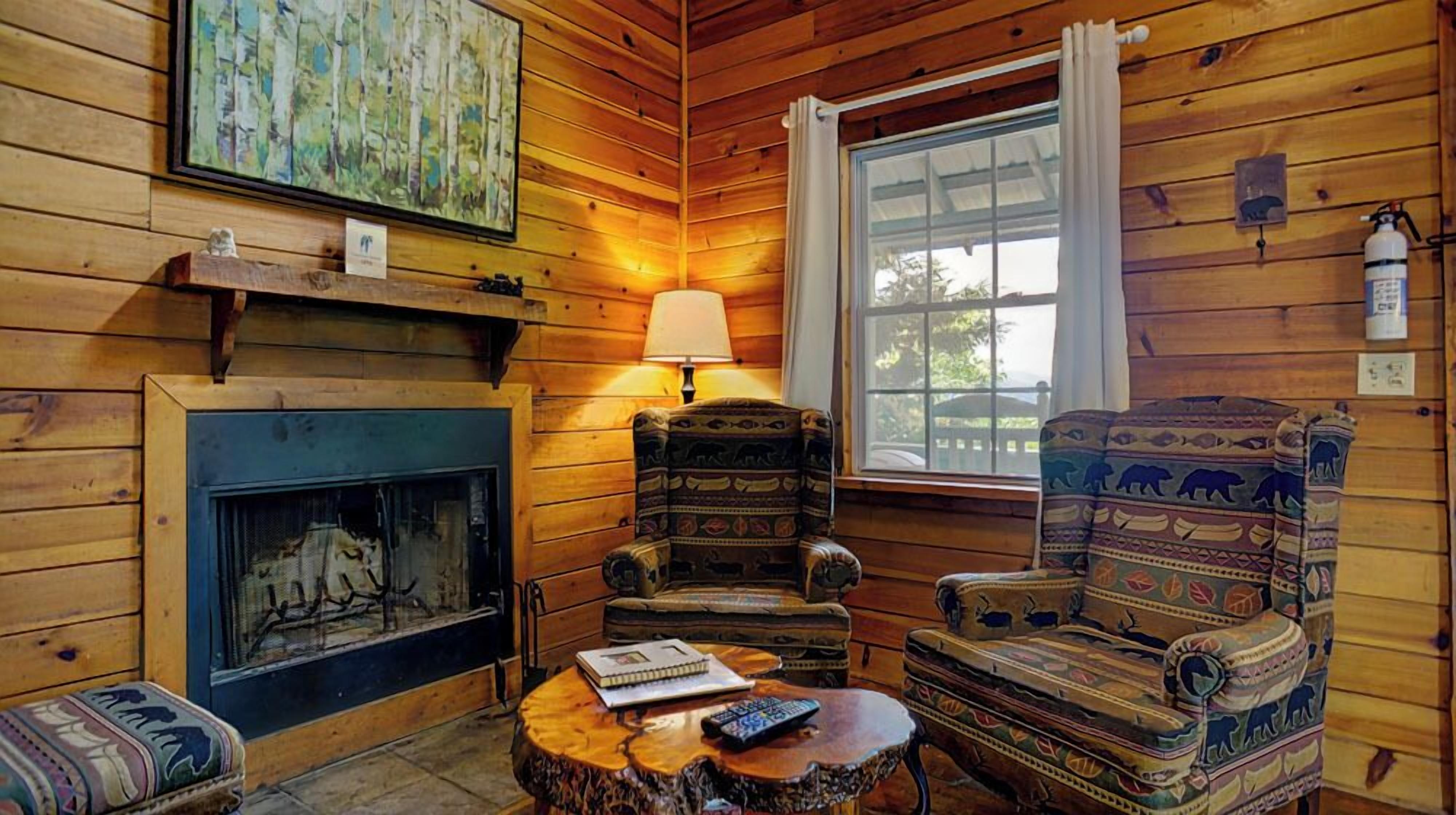 A cozy cabin interior with a fireplace, two patterned armchairs, a round wooden table with a book and remote control, and a large window with curtains.