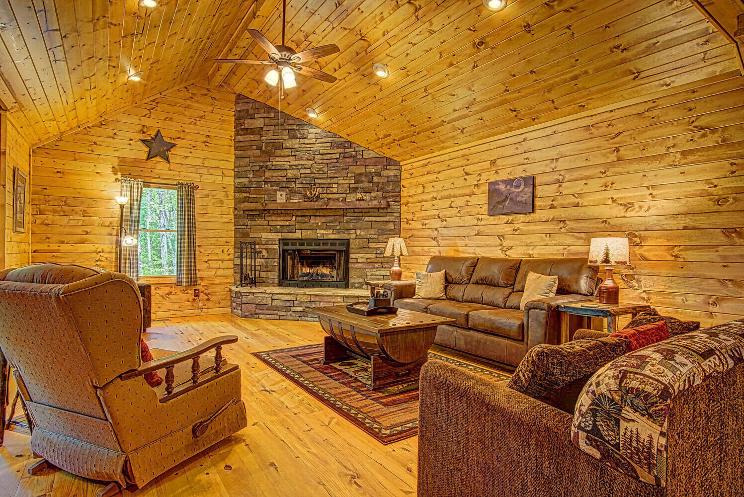 A cozy log cabin interior with a stone fireplace, wooden walls, a ceiling fan, and a comfortable seating area.