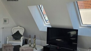 Un salon moderne avec un téléviseur, des poufs, un fauteuil blanc et une cheminée.