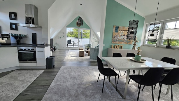 Une cuisine moderne dotée d’un îlot central, d’un coin repas avec une table blanche et des chaises noires, et offrant une vue sur l’extérieur grâce à de grandes fenêtres.