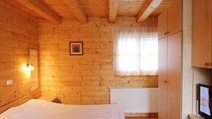 A wooden cabin room with a bed, a window with curtains, a radiator, and a television on a wooden cabinet.