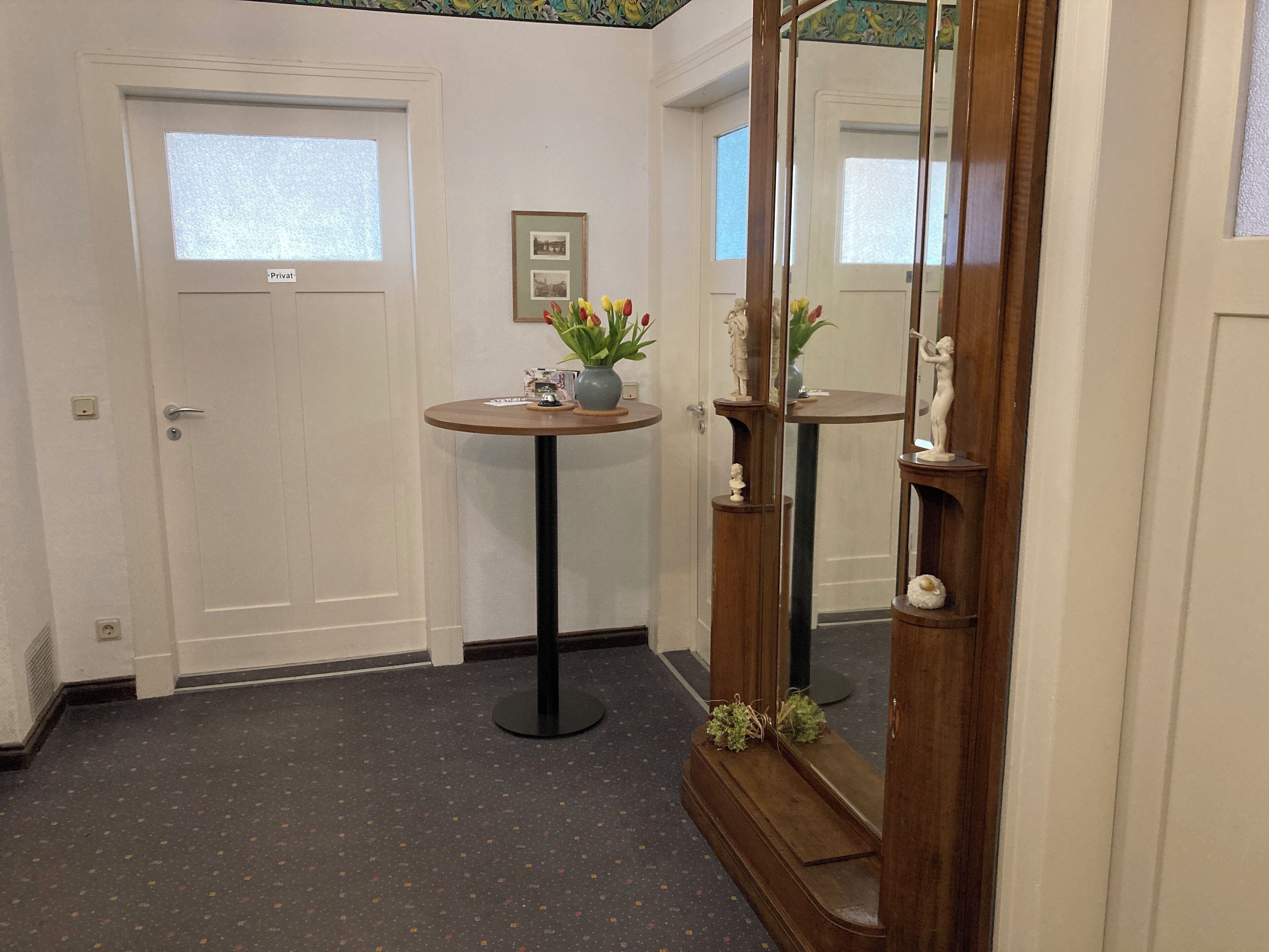 A hallway with a door labeled 'Privat,' a round table with a vase and a small statue, and a mirror reflecting the statue.