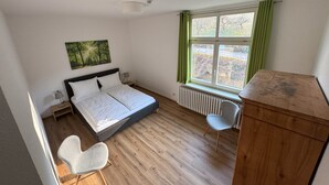 Ein Schlafzimmer mit Bett, Sessel, Heizkörper, Fenster mit Vorhängen und einem Bild an der Wand.