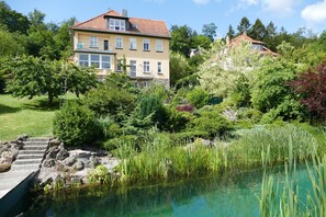 A two-story house with a red-tiled roof, surrounded by lush greenery and a pond. - Modern vacation apartment (Waltershausen)