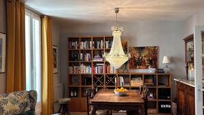 Un salon comprenant une table à manger en bois, un lustre, une bibliothèque et un canapé recouvert d’un tissu à motifs.