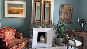 Une pièce avec une cheminée, des œuvres d’art encadrées, un fauteuil rouge recouvert de tissu et un bureau en bois.