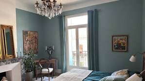 Une chambre à coucher comprenant un lit, un bureau, une chaise, un lustre et une grande fenêtre avec des rideaux.