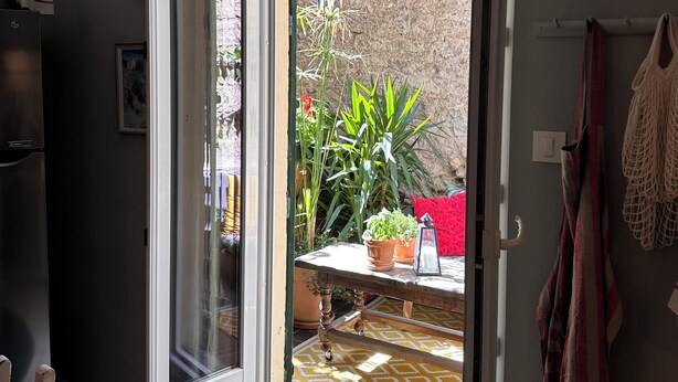 Une pièce dotée d’une porte-fenêtre coulissante qui donne sur une terrasse. On y trouve une chaise en bois, une petite table ornée de plantes en pot et un tapis à motifs rouges et blancs sur le sol.