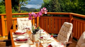 Una terraza de madera con una mesa de comedor para cuatro personas, rodeada de sillas con cojines, con vistas a un paisaje pintoresco con árboles y una masa de agua.