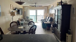 A dining area with a round table, chairs, and a ceiling fan. There is a sofa and a cabinet in the background.