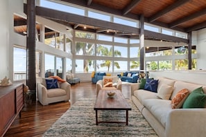 A spacious living room with a high ceiling, large windows, and a view of palm trees. - "Tidepool Getaway" | Oceanfront, A/C, Remodel 2026 (Kahuku)