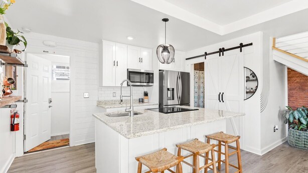 Una cocina moderna con armarios blancos, un refrigerador de acero inoxidable, un microondas y un lavamanos. Hay tres taburetes de madera en la isla.