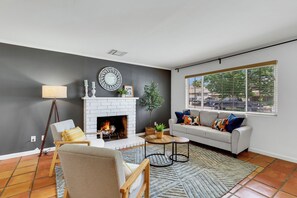 Una sala de estar con chimenea, un sofá, un sillón y un ventanal con persianas.