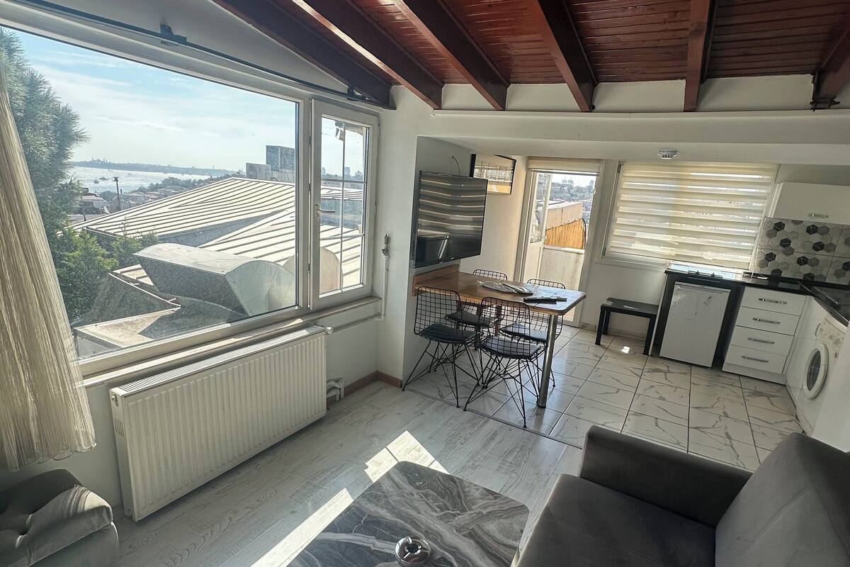 A modern living room with a large window, a sofa, a dining area, and a kitchen.