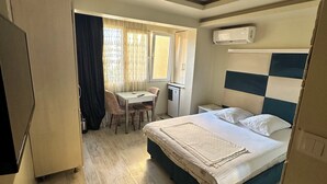 Ein Hotelzimmer mit einem Bett, einem Schreibtisch und einem Stuhl. Es gibt ein Fenster mit Vorhängen, eine Klimaanlage und einen Fernseher.