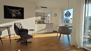 A modern living room with a sofa, a dining table, and chairs. A kitchen is visible in the background.