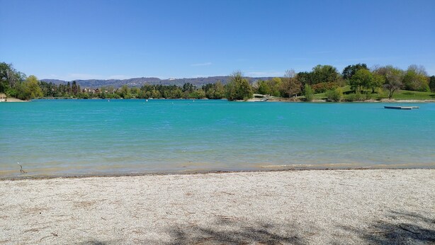 A clear lake with a pebble beach, surrounded by trees and distant hills.
