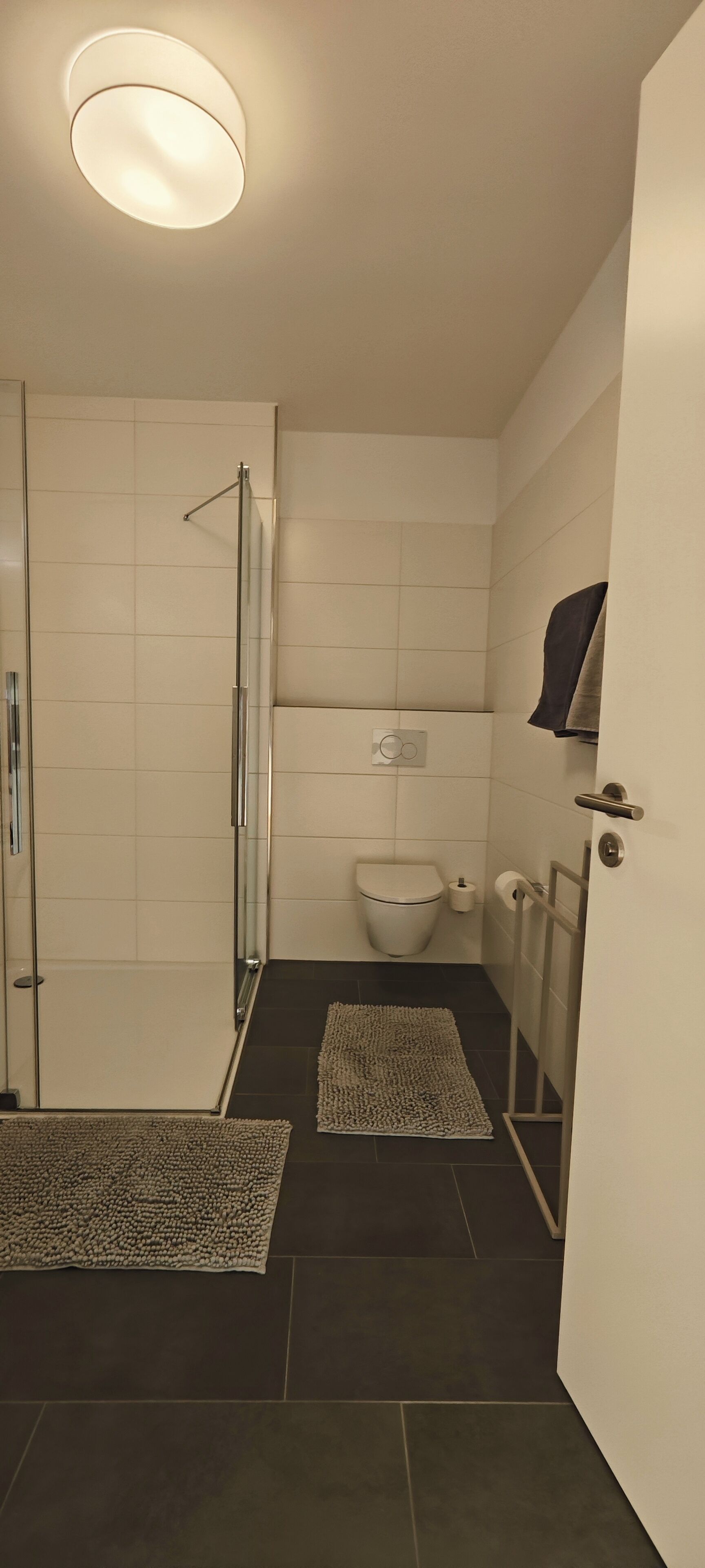Un baño moderno con cabina de ducha de vidrio, azulejos blancos y suelo oscuro.