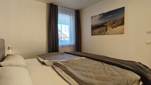 A hotel room with two beds, a large window with curtains, a painting on the wall, and a lamp on the nightstand.