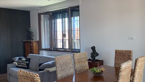 A modern living room with a dining area, featuring a grey sofa, wicker chairs, a wooden dining table, and a large window with blinds. - Cozy apartment 2 steps from Rome finely renovated and equipped (Marino)