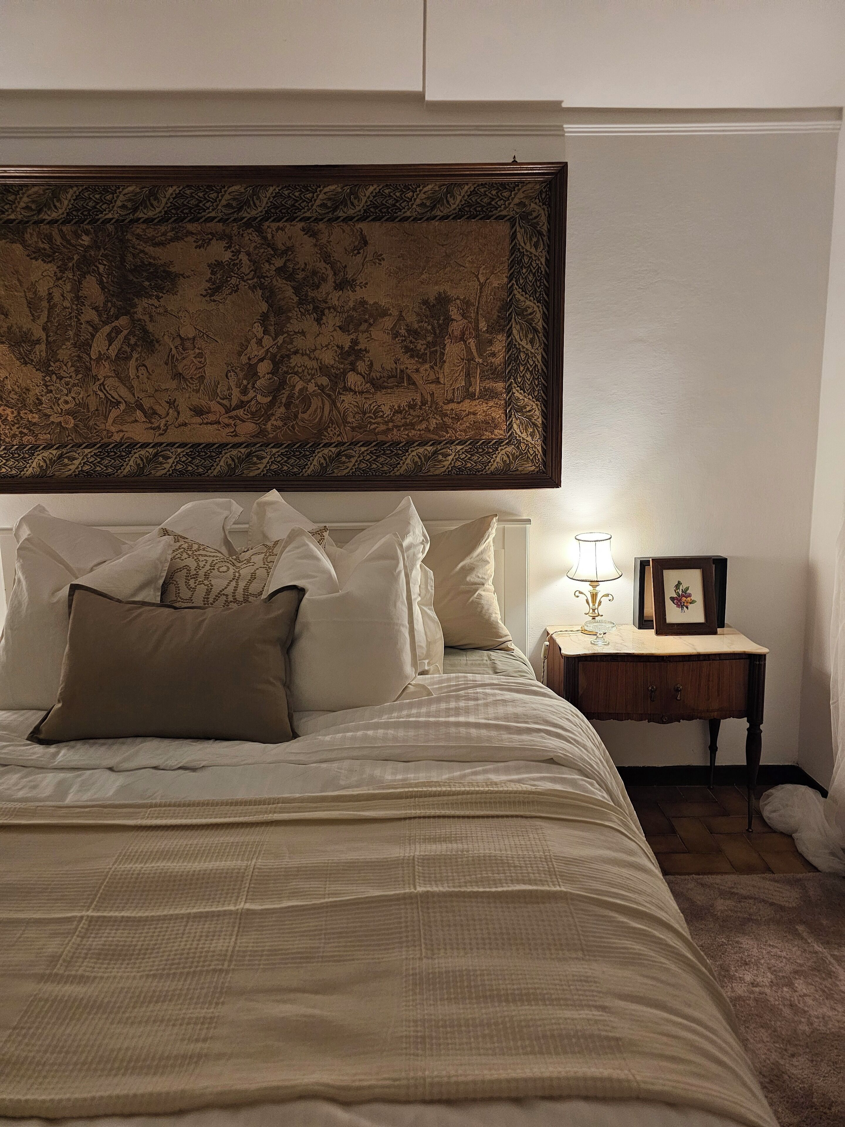 A neatly made bed with a large tapestry above it, a bedside table with a lamp and a picture frame, and a small rug on the floor.