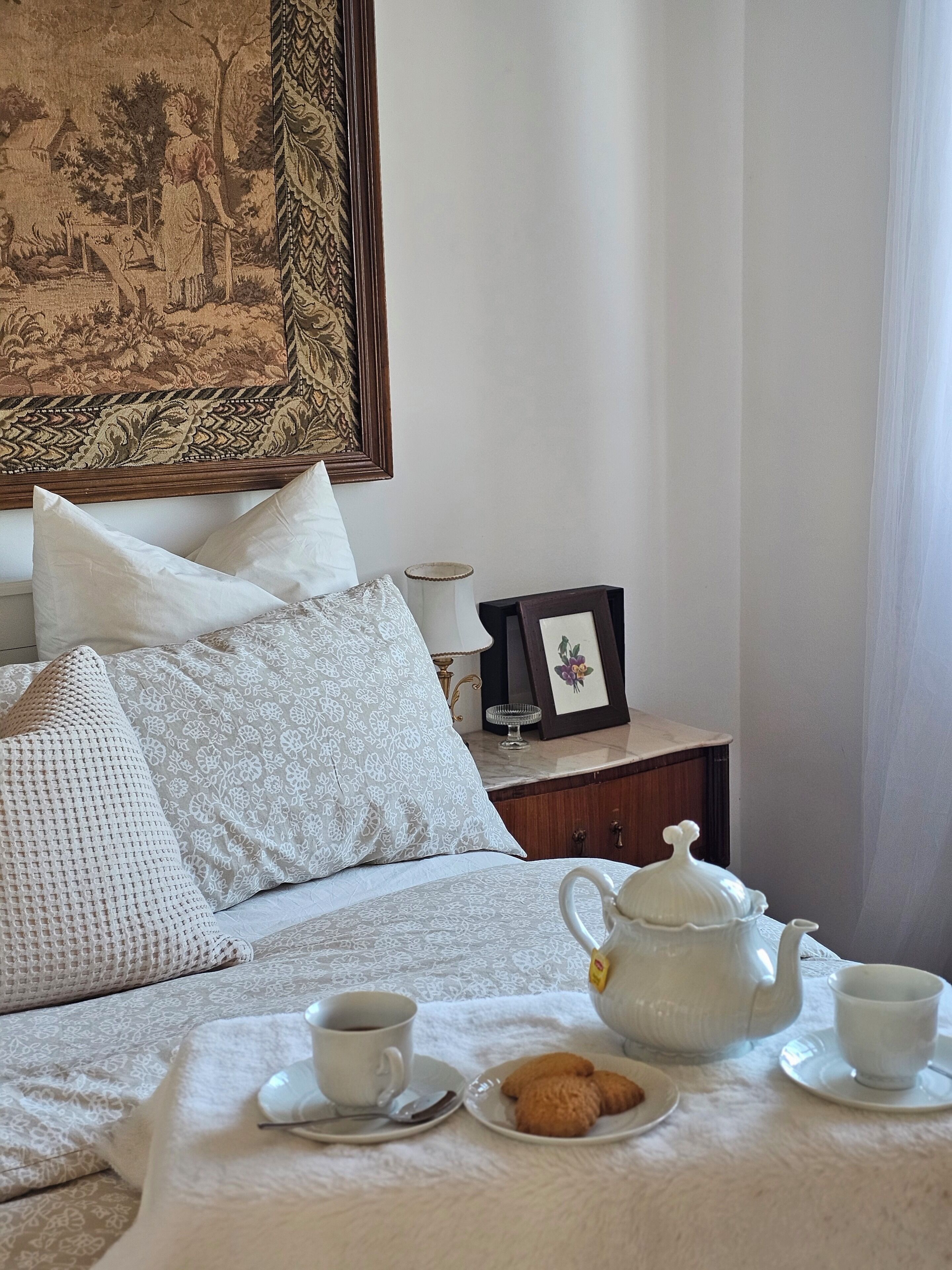 A neatly made bed with white linens and pillows, a bedside table with a lamp and a framed picture, and a tray with a teapot, cups, and cookies on the bed.
