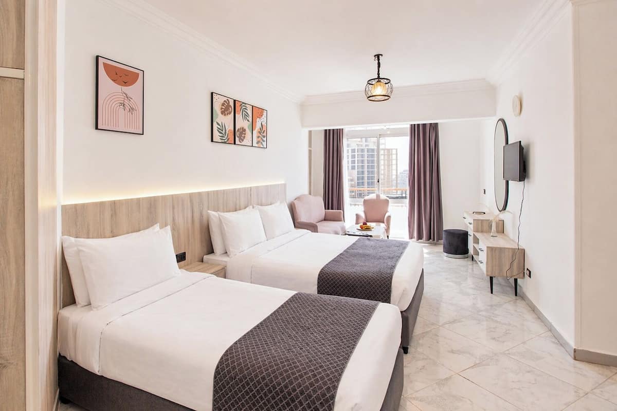 Quarto de hotel com duas camas, uma escrivaninha, uma cadeira, televisão e uma janela com cortinas.