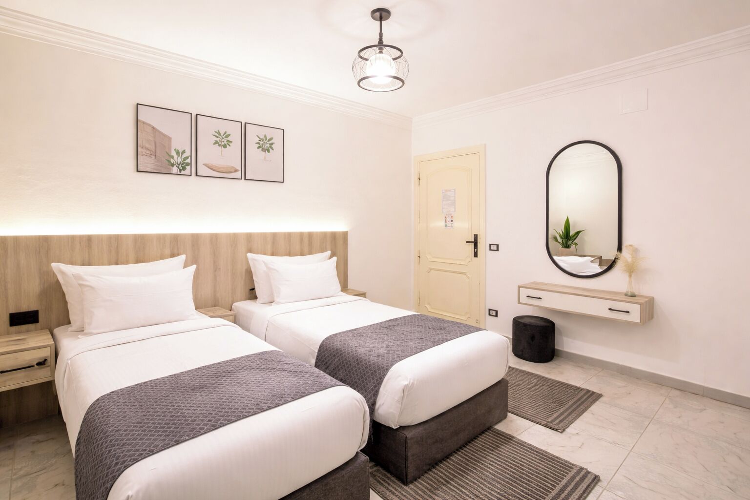 Habitación de hotel con dos camas, una mesita de noche, un espejo, una planta pequeña y una puerta.