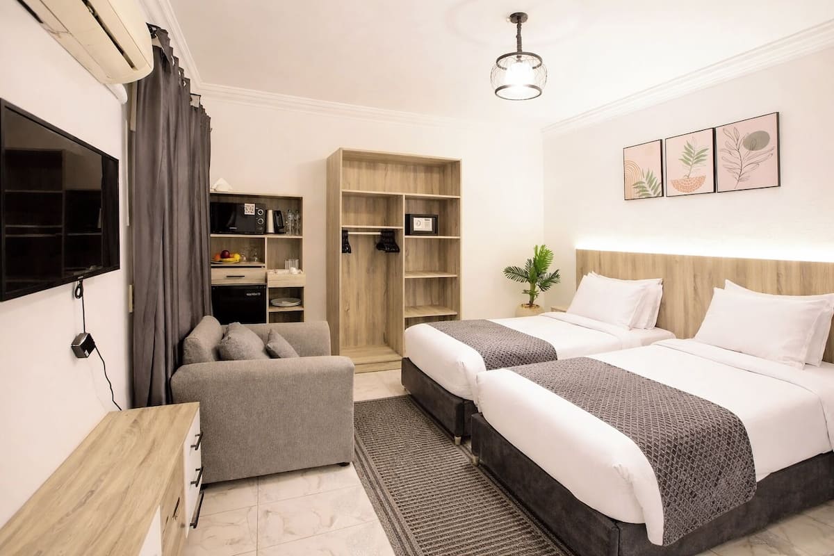 Quarto de hotel com duas camas, um sofá, uma televisão, um guarda-roupa e uma pequena planta.