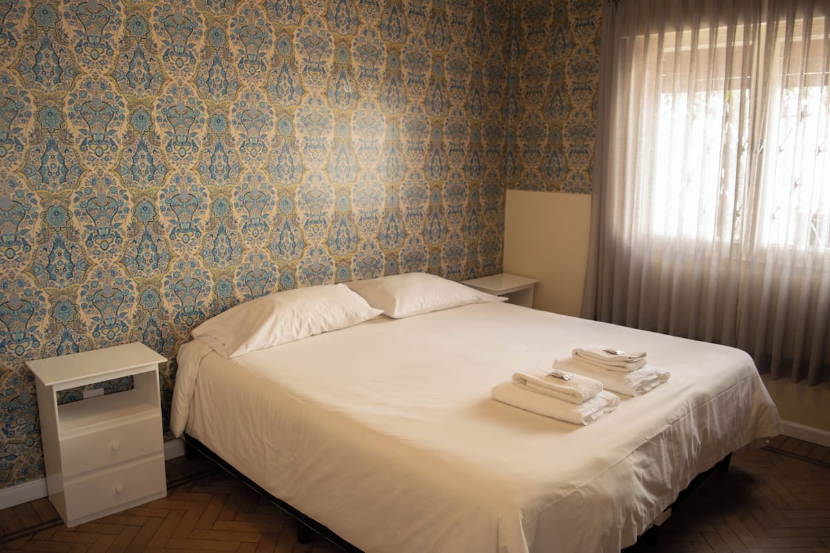A neatly made bed with white linens and pillows, a bedside table with a lamp, and a window with sheer curtains.