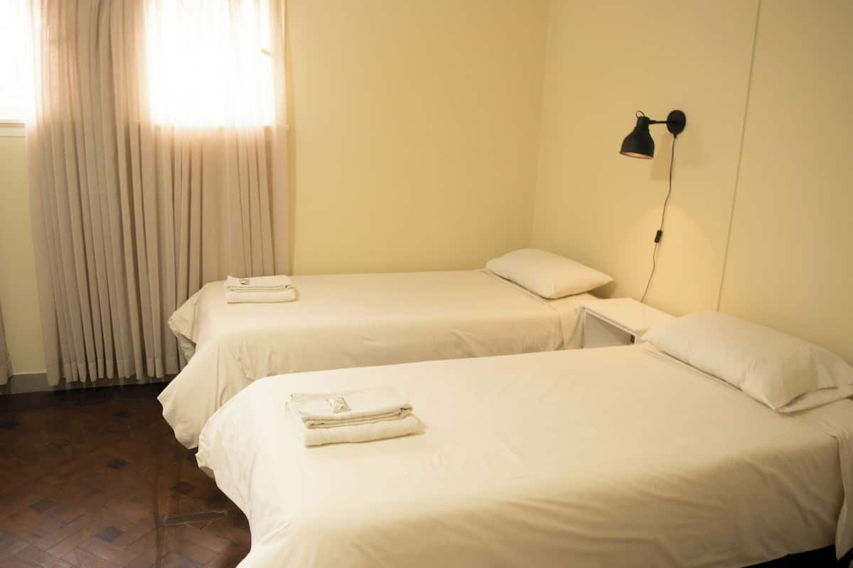 A hotel room with two single beds, a bedside table with a lamp, and a window with curtains.