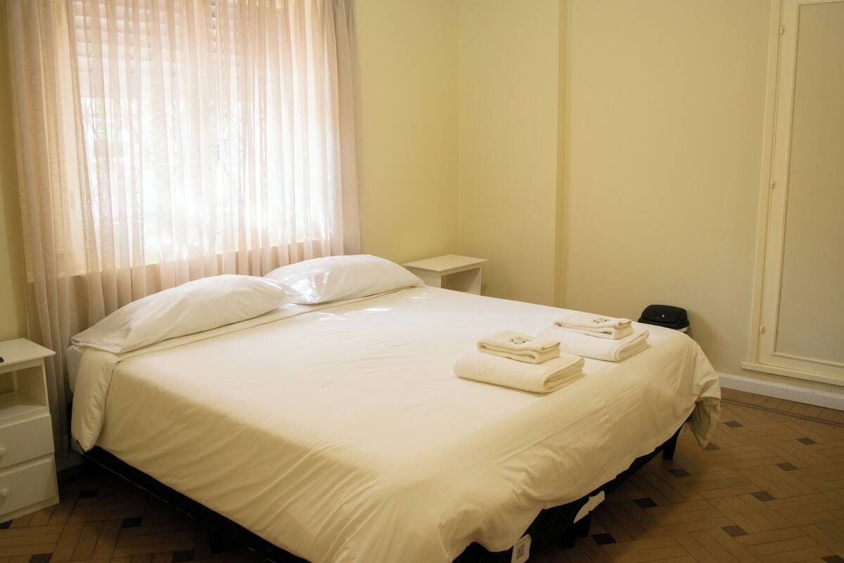 A neatly made bed with pillows and folded towels, a bedside table, and a window with sheer curtains.