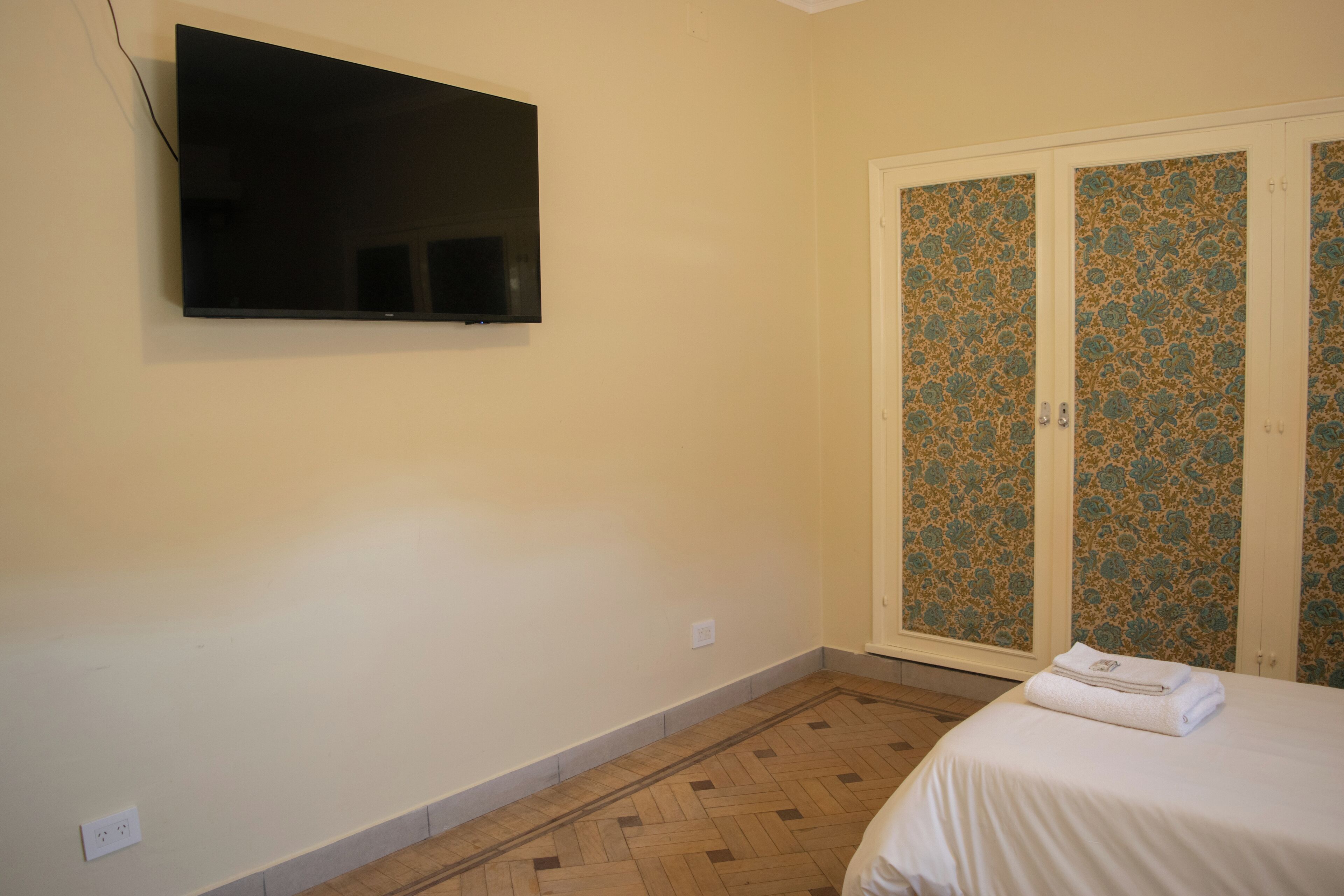 A bedroom with a flat-screen TV mounted on the wall, a bed with a white bedspread, and a wooden herringbone floor.