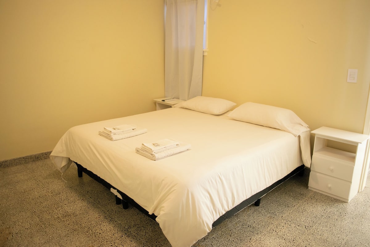 A neatly made bed with white linens and two folded towels, a bedside table with a lamp, and a nightstand with a lamp.