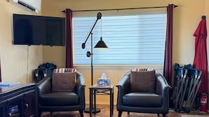 A living room with two leather armchairs, a small side table, a floor lamp, a flat-screen TV, and a window with blinds.