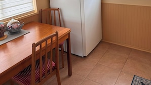 A compact kitchen with a white refrigerator, wooden dining table, chairs, and a window with blinds.