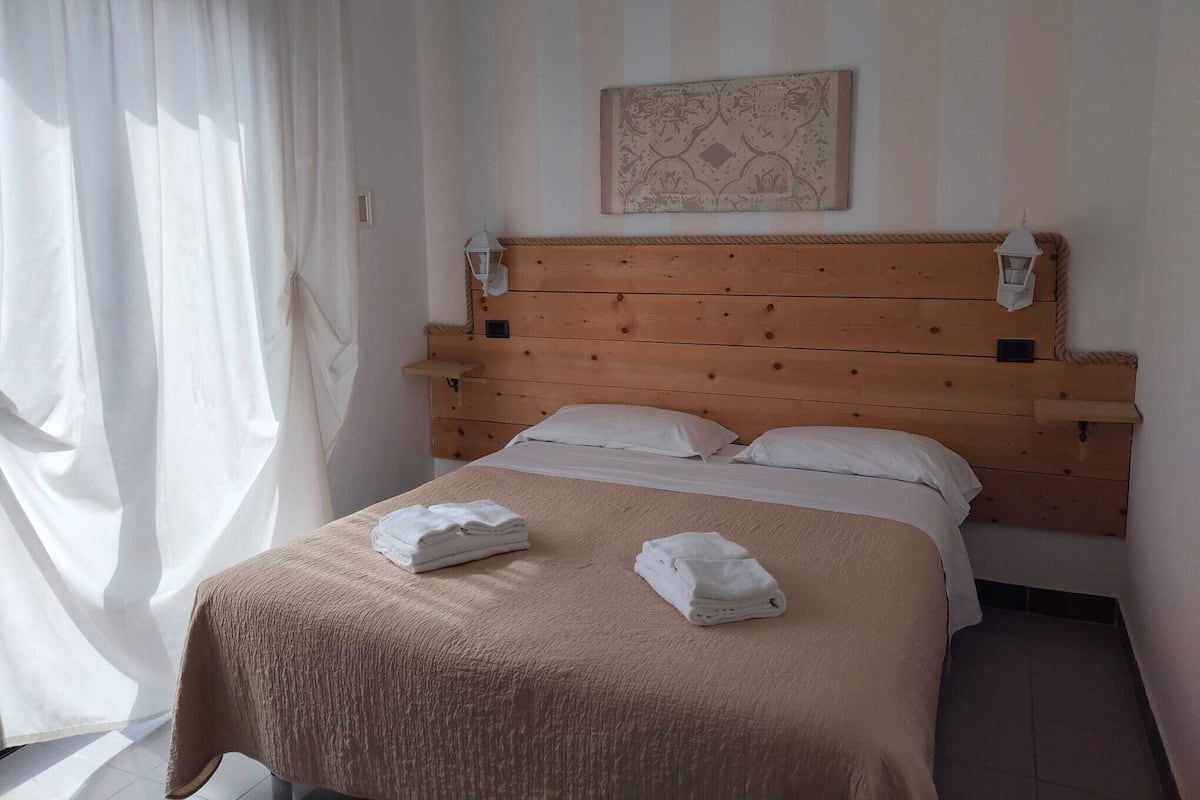 Een hotelkamer met een houten hoofdeinde, een bed met witte lakens en een raam met dunne gordijnen.